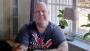Bilden visar en man med skallig hjässa och skägg, sittande vid ett fönster. Han bär en mörkblå t-shirt med röd-vit text/logotyp. Mannen har tatueringar på armen och ser rakt in i kameran med ett allvarligt ansiktsuttryck. I bakgrunden syns ett fönster med ljusa gardiner, en krukväxt med stora gröna blad och en grå bordslampa på fönsterbrädan. Genom fönstret kan man skymta en utomhusmiljö med ljusa strukturer, möjligen en altan eller trädäck. Miljön ger intryck av ett hemtrevligt vardagsrum eller liknande utrymme med naturligt ljus som strömmar in genom fönstret.