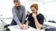 Sebastian Lindgren sitter ner i skolbänken tillsammans med läraren Peter Muskos, som står upp bredvid honom.