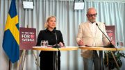 Bilden visar en presskonferens med Magdalena Andersson och Johan Lindholm som står vid varsitt podium. Till vänster syns en Magdalena i svart dräkt och till höger Johan i ljus kostym med glasögon. Bakom dem finns en svensk flagga (blå med gult kors) och skyltar med texten "NY RIKTNING FÖR SVERIGE" och "KONGRESS 2025" tillsammans med en socialdemokratisk ros-symbol. Bakgrunden består av ljusa draperier och det finns belysningsutrustning i taket. På podiumbordet framför varje person finns ett vattenglas och en mikrofon.